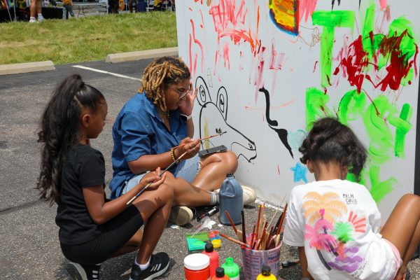 An adult and two children sitting on the ground paint a mural on a white wall together.